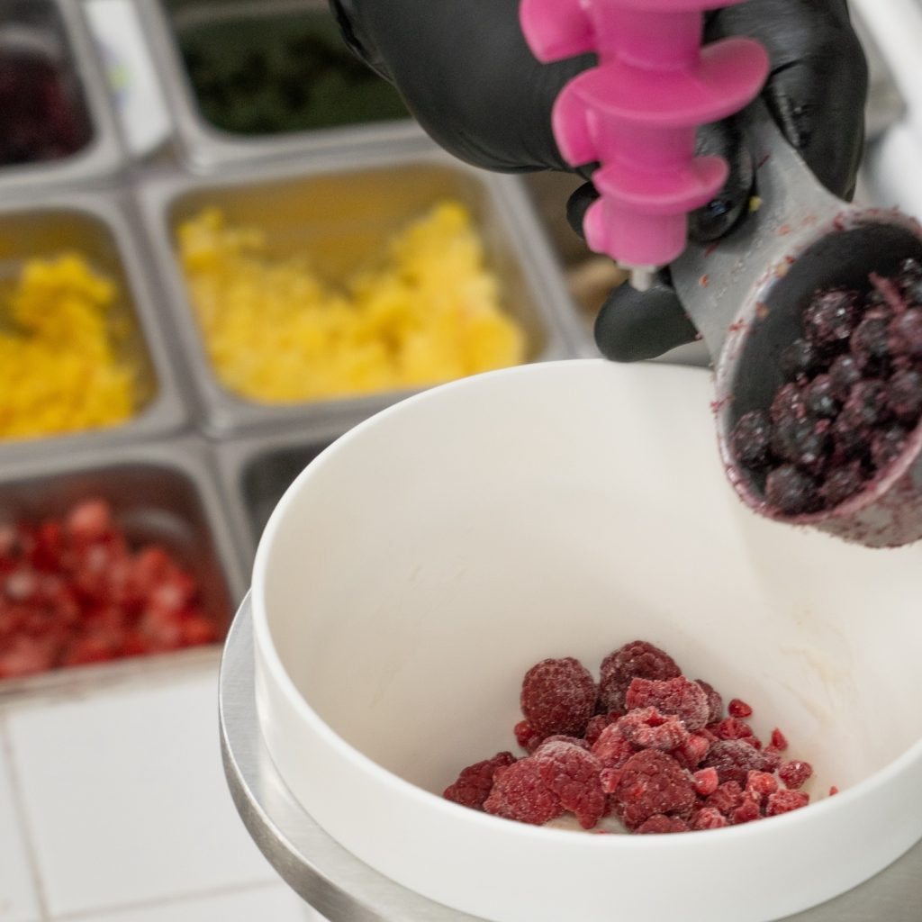 Black-gloved hand scooping frozen mixed berries into a white bowl, with containers of colourful frozen fruits in the background.
